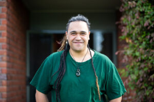 A person with long dreadlocks and a greenstone pendant smiling warmly in a doorway