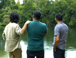 Three people standing together by a lake, two embracing with arms around each other, viewed from behind